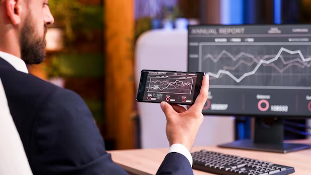 A man sits in front of a computer and holds a phone in his hands, which shows a graphic